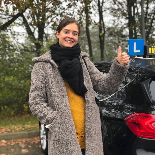 Leerling met grijze jas en opgestoken duim naast de Meteor-lesauto.