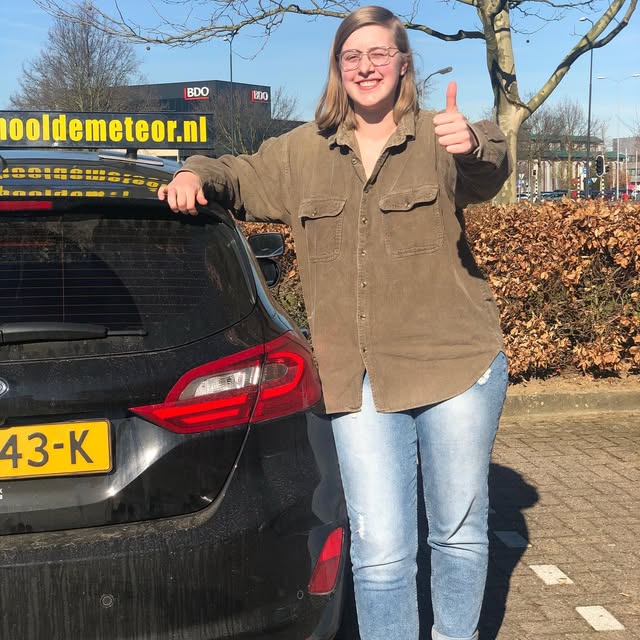 Leerling met bril en opgestoken duim achter de Meteor-lesauto.